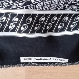 Puede incluir: Un textil batik en blanco y negro con intrincados patrones geométricos y tipo paisley. La tela presenta un borde con el texto "BATIK Tradisional BY DORA". Los bordes no están acabados.