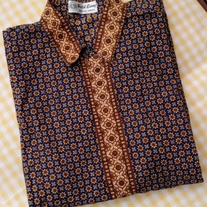Könnte beinhalten: Ein langärmeliges Hemd mit Knopfleiste aus dunkelblauem Stoff mit einem sich wiederholenden beigen und braunen floralen Batikmuster. Der Kragen und die Knopfleiste zeigen ein kontrastierendes beiges und braunes geometrisches Batikdesign. Ein Etikett zeigt Batik Enny.