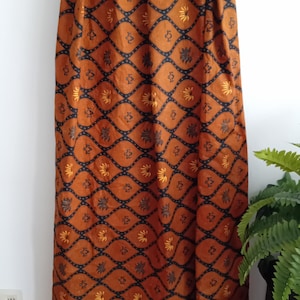 Könnte beinhalten: Ein langer, gemusterter Stoff in Braun-, Schwarz- und Gelbtönen ist über einem Holzgestell drapiert. Der Stoff weist ein sich wiederholendes geometrisches Design mit floralen und Punktmotiven auf. Die Gesamtästhetik ist vintage und dekorativ.