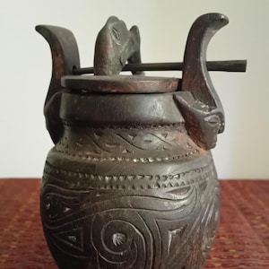 May include: Dark brown, hand-carved wooden vessel with a lid and handles. The vessel features intricate carvings and a detailed design. The handles are shaped like stylized figures. The lid has a small animal carving. The vessel is likely used for storage.