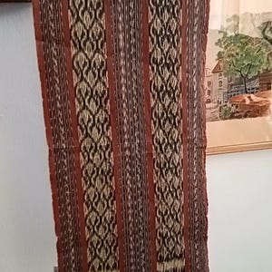 Op de afbeelding: Een rechthoekige geweven textielloper met verticale strepen in tinten bruin, zwart en crème. De stof toont een herhalend ingewikkeld geometrisch patroon binnen de bredere banen en is voorzien van franjes langs de onderrand.