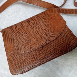 May include: A brown, rectangular crossbody bag with a flap closure. The bag has a textured, reptile-like pattern and a long, thin strap. The bag is made of a leather-like material.