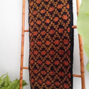 Op de afbeelding: Een zwarte textiel met een ingewikkeld patroon van oranje en rode bloemmotieven. De stof is over een bamboeladder gedrapeerd, met een franje aan de onderkant. Waarschijnlijk een sjaal of omslagdoek.