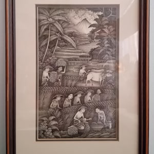 Op de afbeelding: Ingelijste zwart-wit kunstwerk dat een landelijke scène toont met figuren die gewassen oogsten, een koe en palmbomen. Het kunstwerk is geplaatst in een donkerbruine houten lijst met een crèmekleurige passe-partout.