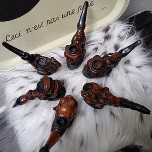 Può includere: Sei pipe in legno decorative con intagli intricati e steli neri. Le pipe sono disposte su una superficie bianca e soffice. Il testo "Ceci n'est pas une pipe" è visibile sullo sfondo.