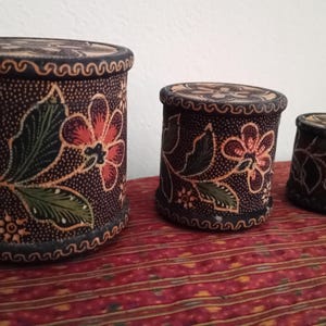 Pode incluir: Conjunto de três caixas cilíndricas decorativas com tampas. As caixas apresentam um padrão floral em vermelho, verde e laranja sobre um fundo castanho escuro. As tampas e as bordas têm uma borda decorativa.