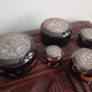 May include: A set of four round containers with dark brown and black mottled patterns. Each lid features a circular silver-toned metal inlay with an intricate embossed floral design. The containers vary in size, arranged in a cluster.