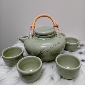 Puede incluir: Un juego de té de cerámica en color verde salvia que incluye una tetera con tapa y asa trenzada curva, junto con cuatro tazas pequeñas a juego. Cada pieza presenta un acabado de esmalte craquelado y un delicado diseño de hojas oscuras.