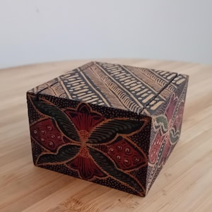 Könnte beinhalten: Eine quadratische Holzschachtel mit dunkelbrauner und schwarzer Grundfarbe und einem handgemalten Blumenmuster in Rot-, Grün- und Cremetönen. Der Deckel und die Seiten sind mit komplizierten, sich wiederholenden Motiven und feinen Punkttexturen verziert.