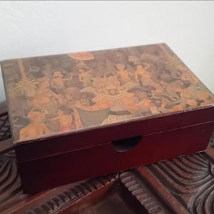 Può includere: Una scatola rettangolare in legno marrone scuro con coperchio incernierato. La superficie superiore presenta un'illustrazione dettagliata e multicolore di diverse figure in abiti tradizionali, tra cui un personaggio centrale con maschera.