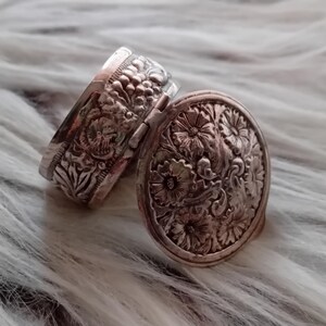 Könnte beinhalten: Zwei antike silberne Medaillons mit aufwendigen floralen Designs. Eines ist geöffnet und zeigt das detaillierte Innere. Das andere ist geschlossen und zeigt ein passendes Blumenmuster. Die Medaillons sind vor einem weichen, strukturierten Hintergrund platziert.