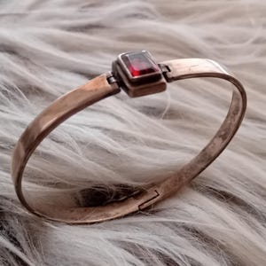 Op de afbeelding: Een zilverkleurige armband met een rechthoekige, dieprode edelsteen in het midden. De armband heeft een scharnierende sluiting en een eenvoudig ontwerp. De edelsteen is ongeveer 1,3 cm lang.
