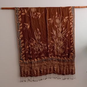 Op de afbeelding: Een bruin textiel met een bloemenpatroon en franjes, gedrapeerd over een houten stok. Het ontwerp toont gestileerde bloemen en bladeren in een lichtere bruine kleur, met een donkerdere bruine rand en franjes. Waarschijnlijk een sjaal of omslagdoek.