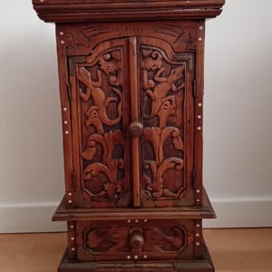 Könnte beinhalten: Ein kleiner, aufwendig geschnitzter Holzschrank mit zwei Türen und einer Schublade. Der Schrank weist detaillierte Reliefschnitzereien von stilisierten Figuren und floralen Mustern auf. Das Holz hat eine warme braune Farbe und ist mit kleinen, dekorativen Akzenten verziert.