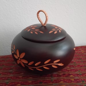 May include: A dark brown, round wooden container with a lid. The container is decorated with carved floral and leaf patterns in a lighter brown colour. It has a loop handle on top.