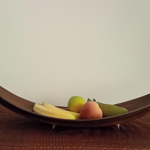 Peut inclure: Un bol à fruits en bois de forme incurvée, contenant des bananes, une pomme verte, une pêche et une poire. Le bol est marron et repose sur de petits pieds en métal. Le bol à fruits est sur une surface marron.
