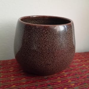 Peut inclure: Une tasse en céramique marron tachetée avec une large ouverture. La tasse a une forme arrondie et repose sur une surface à motifs rouges et dorés. La tasse est probablement utilisée pour boire des boissons.