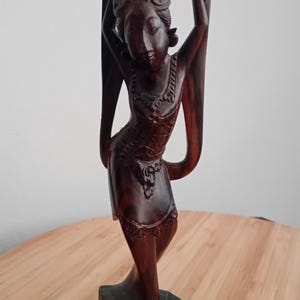 Op de afbeelding: Een donkerbruin handgesneden houten beeldje van een staande figuur met de armen omhoog in een gracieuze houding. Het beeldhouwwerk is voorzien van gedetailleerde versieringen op het hoofddeksel en de kleding, met een gladde, gepolijste afwerking.