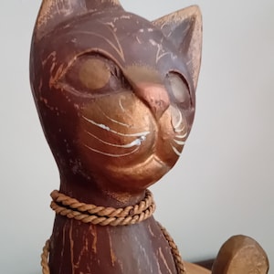 Könnte beinhalten: Eine geschnitzte Katzenfigur aus Holz mit dunkelbraunem Finish und goldfarbenen Akzenten an Ohren, Gesicht und Schwanz. Die Katze trägt ein dekoratives, beigefarbenes Seilhalsband um den Hals. Die Holzmaserung ist auf der gesamten Oberfläche sichtbar.