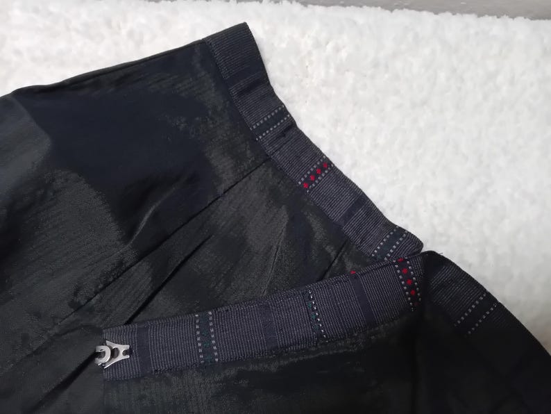 May include: Black skirt with a plaid waistband and a black fabric belt with a silver buckle. The skirt has a pleated design.