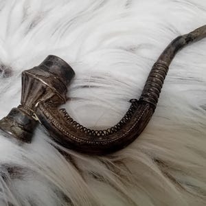 May include: An antique silver-toned smoking pipe with a curved stem and ornate detailing. The bowl has a decorative pattern, and the stem is wrapped with a textured design. The pipe rests on a fluffy, white surface.
