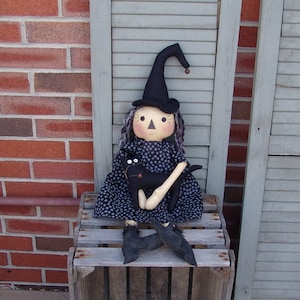 May include: A handmade witch doll wearing a black hat and a blue and white polka dot dress. She is holding a black cat in her arms and sitting on a wooden crate.