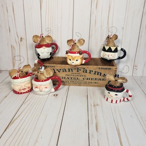 May include: Set of six ceramic Christmas ornaments shaped like mugs with a mouse peeking out of the top. Each mug features a different holiday character: Santa Claus, a snowman, a reindeer, a penguin, and two more Santas. The mugs are decorated with red, white, and black accents.