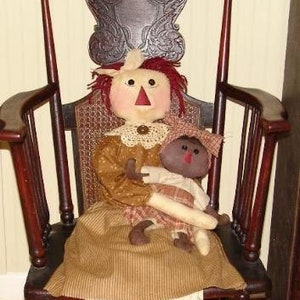 Könnte beinhalten: Eine Stoffpuppe mit roten Haaren und einem braunen Kleid sitzt in einem hölzernen Schaukelstuhl. Die Puppe hält eine kleinere Stoffpuppe mit brauner Haut und einem karierten Kleid.