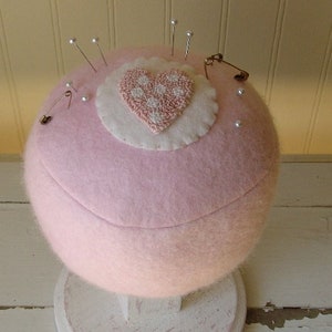 May include: A pink felt pincushion with a white heart-shaped applique with pink polka dots. The pincushion is sitting on a white pedestal.