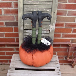 May include: A handmade witch doll with black boots and green legs, sitting on a pumpkin. The doll is wearing a black tulle skirt and has a tag that reads "Black Hat Secrets" with a witch silhouette.