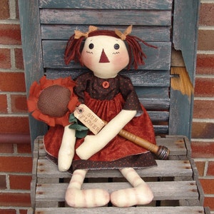 May include: A rag doll with red hair, wearing a brown and black dress, holding a sunflower. The doll is sitting on a wooden crate with the words "Sun Flower" on a tag.