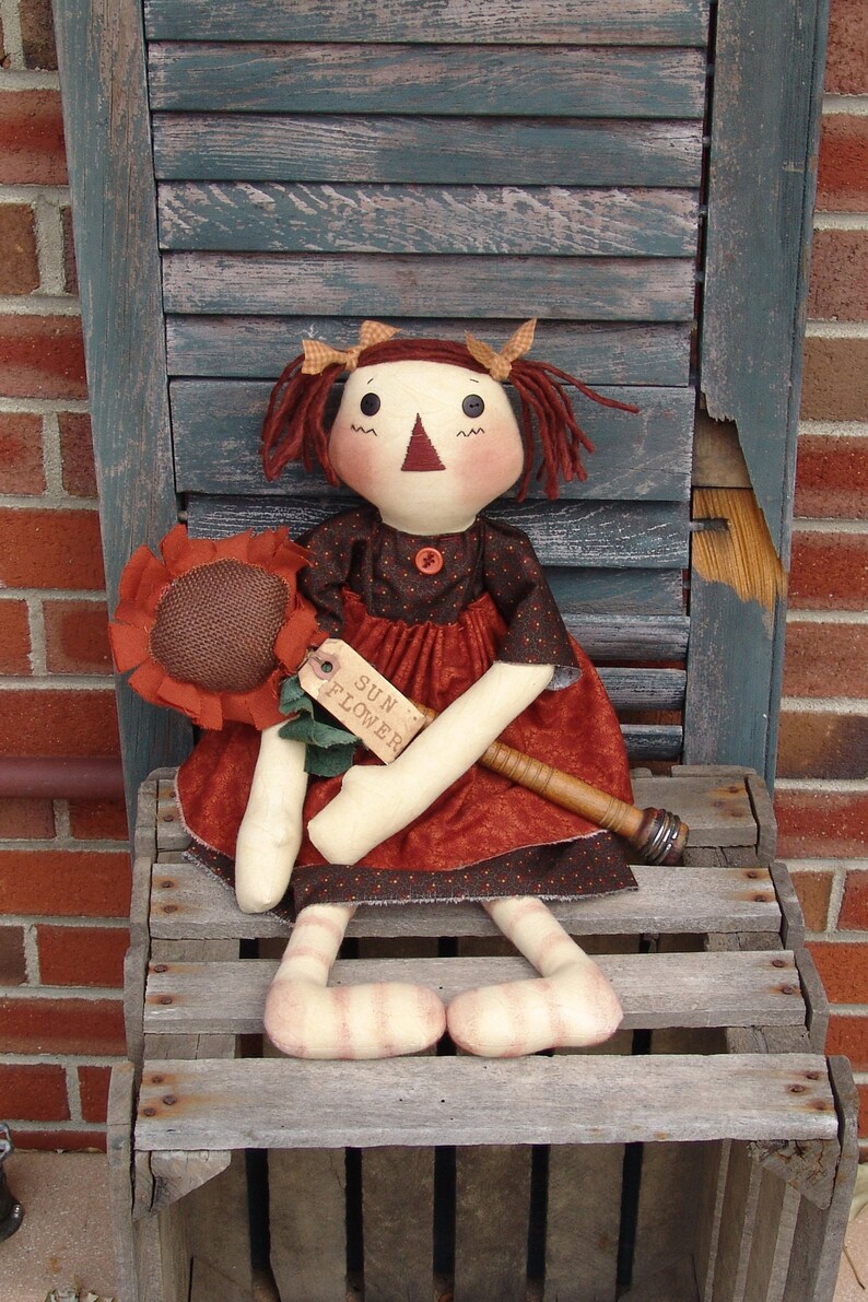 May include: A rag doll with red hair, wearing a brown and black dress, holding a sunflower. The doll is sitting on a wooden crate with the words "Sun Flower" on a tag.