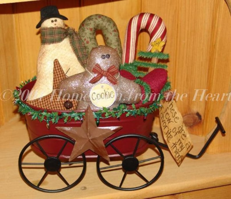 May include: A red wagon filled with Christmas decorations, including a snowman, gingerbread man, candy cane, and a star. The wagon has black wheels and is decorated with green garland. The text "These are a few of my favorite things" is written on a tag attached to the wagon.