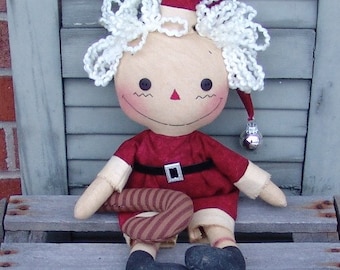 May include: A handmade rag doll dressed as Santa Claus. The doll is wearing a red and white Santa suit with a black belt and a white fur trim. The doll is holding a candy cane in its hand and has a white yarn wig. The doll is sitting on a wooden bench.