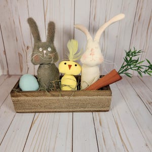 May include: A rustic wooden box filled with Easter decorations. It contains a gray bunny, a yellow chick with a feather, and a white bunny with a carrot. An egg is also in the box. The box is lined with burlap.