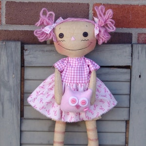 May include: A pink rag doll with pink yarn hair, wearing a pink and white gingham dress with pink ribbon accents. The doll is holding a pink felt owl with white eyes.