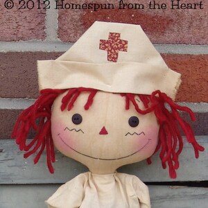 May include: A handmade doll with red yarn hair and a nurse's cap featuring a cross design. The doll has button eyes, a stitched smile, and rosy cheeks. The doll is wearing a cream-colored outfit. The text "&copy; 2012 Homespun from the Heart" is visible.