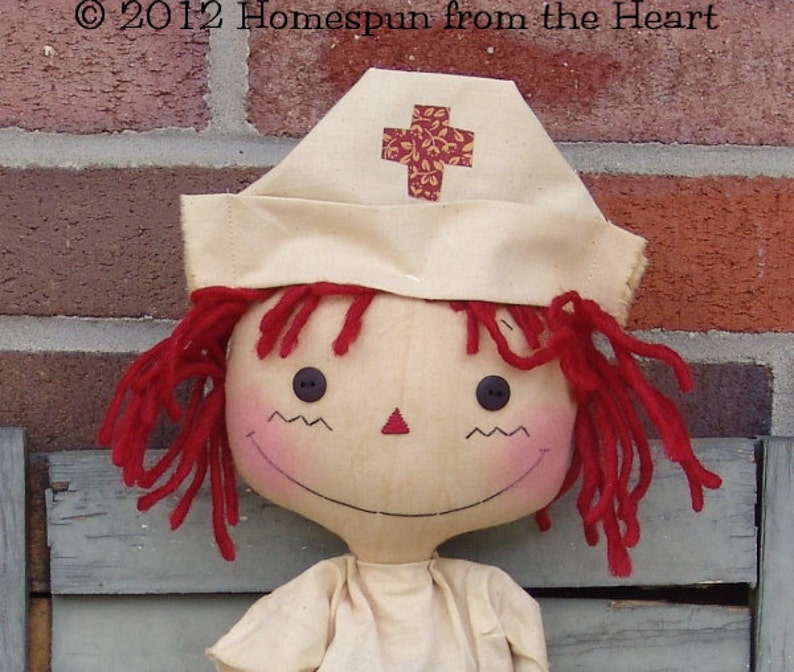 May include: A handmade doll with red yarn hair and a nurse's cap featuring a cross design. The doll has button eyes, a stitched smile, and rosy cheeks. The doll is wearing a cream-colored outfit. The text "&copy; 2012 Homespun from the Heart" is visible.