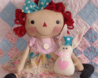 May include: A handcrafted rag doll with red yarn hair, a blue polka dot bow, and a pink gingham dress. Next to it is a white bunny doll wearing a blue polka dot top hat and a pink heart. Both dolls are on a patchwork quilt.