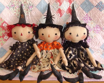 May include: Three handmade rag dolls dressed as witches. Each doll has a black witch hat with a star or spider detail. The dolls are wearing black and white dresses with different patterns. The dolls are sitting on a pink and white quilt.