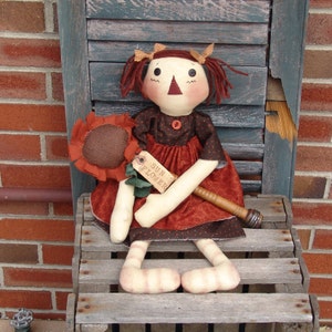 May include: A handmade rag doll with red hair, wearing a brown and red dress with white polka dots. The doll is holding a sunflower and a wooden stick with a tag that says "Sun Flower".