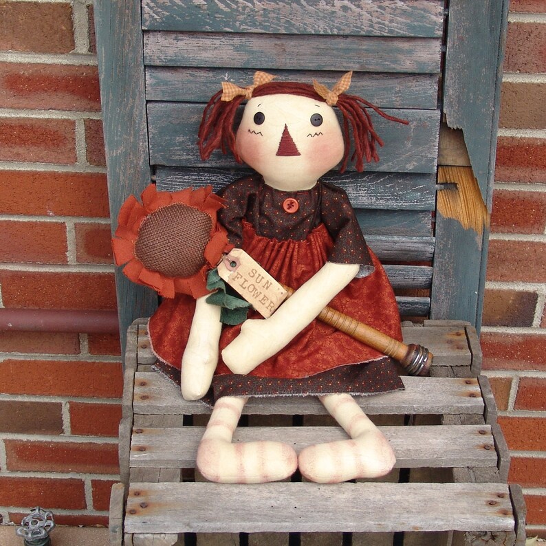 May include: A handmade rag doll with red hair, wearing a brown and red dress with white polka dots. The doll is holding a sunflower and a wooden stick with a tag that says "Sun Flower".
