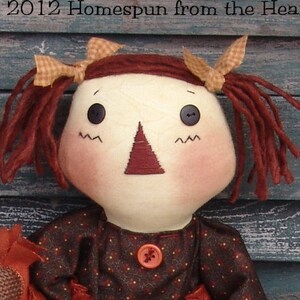 May include: A rag doll with red yarn hair, brown eyes, and a red nose. She is wearing a brown dress with white polka dots and a red button. The doll is holding a brown fabric object.