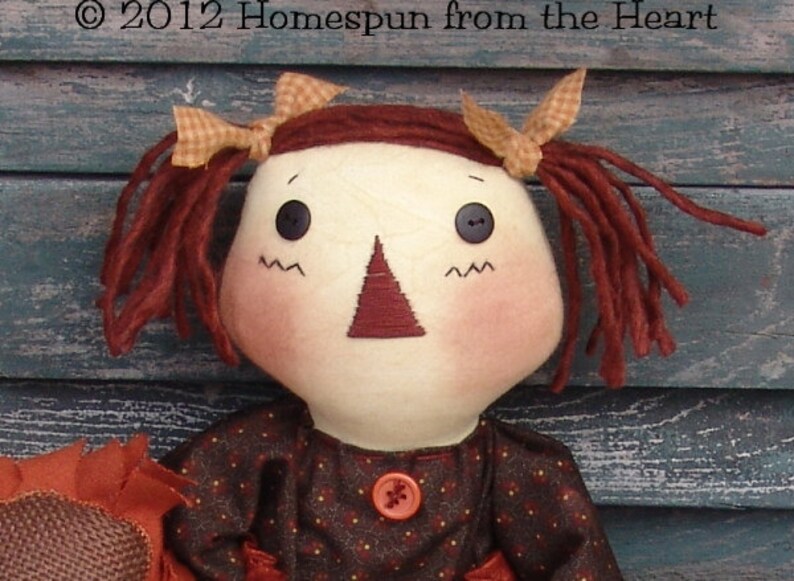 May include: A rag doll with red yarn hair, brown eyes, and a red nose. She is wearing a brown dress with white polka dots and a red button. The doll is holding a brown fabric object.
