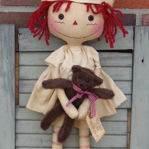 May include: A handmade doll dressed as a nurse, wearing a cream-colored cap with a red cross. The doll has red yarn hair, rosy cheeks, and holds a brown teddy bear with a pink ribbon. A tag with text hangs from the bear.