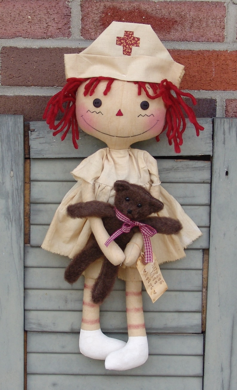 May include: A handmade doll dressed as a nurse, wearing a cream-colored cap with a red cross. The doll has red yarn hair, rosy cheeks, and holds a brown teddy bear with a pink ribbon. A tag with text hangs from the bear.