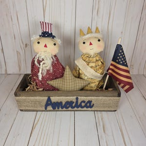 May include: A wooden box with burlap trim contains two fabric dolls, a star, and a small American flag. One doll wears a red and white star-patterned outfit and a top hat. The other doll wears a gold-coloured outfit and a crown. The word "America" is on the box.