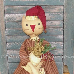 May include: A handmade rag doll cat wearing a red Santa hat and a brown and white striped dress. The cat is holding a small pot with a sprig of greenery and a candy cane.