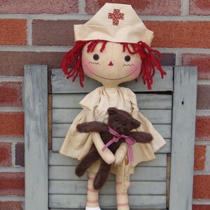May include: A handmade doll dressed as a nurse, with red yarn hair and a matching hat with a cross emblem. The doll is holding a small brown teddy bear with a pink ribbon. The doll is standing in front of a brick wall.