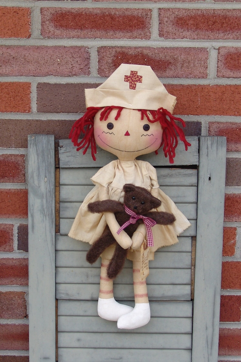 May include: A handmade doll dressed as a nurse, with red yarn hair and a matching hat with a cross emblem. The doll is holding a small brown teddy bear with a pink ribbon. The doll is standing in front of a brick wall.